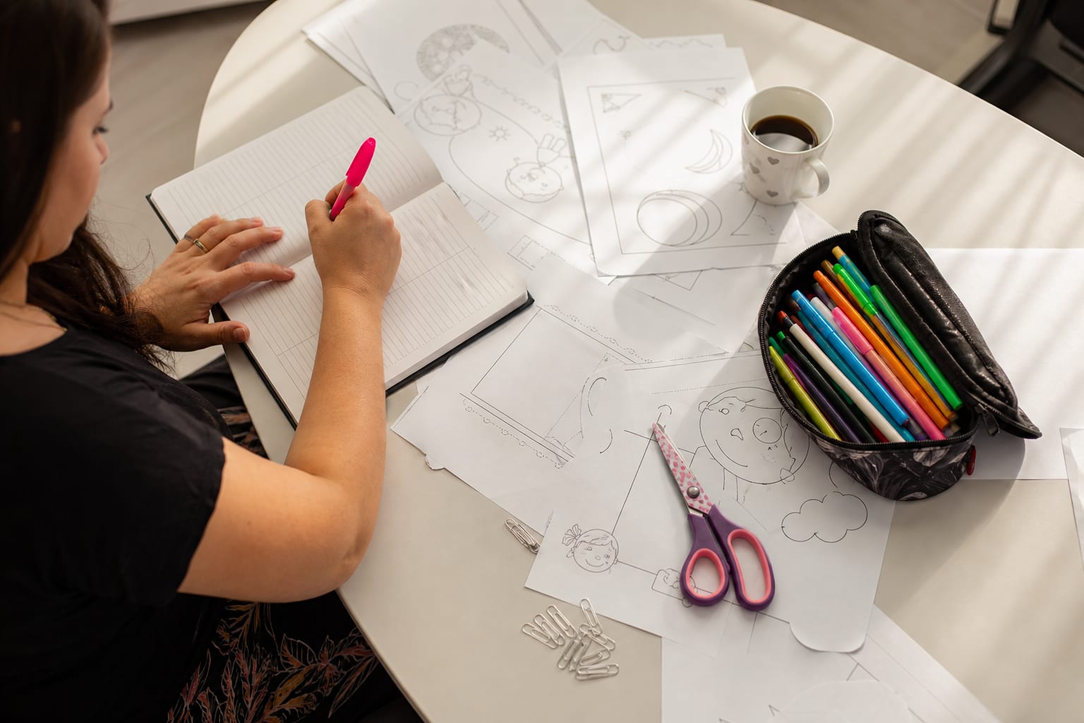 Mesa de professora vista de cima, com folhas de atividade impressas em preto e branco, caderno de planejamento, canetinhas e clipes